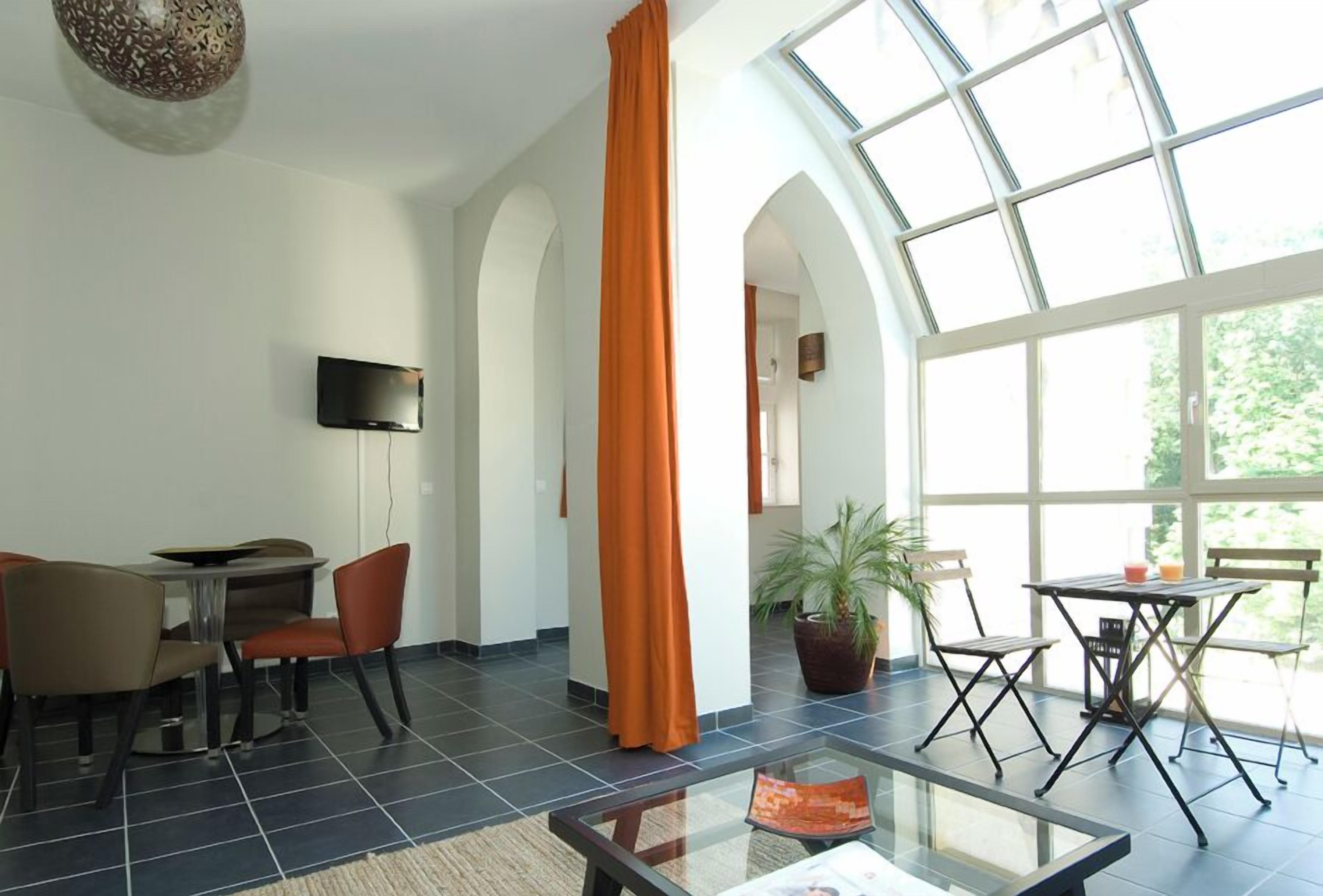 A spacious room with a large window, a flat-screen TV mounted on the wall, and a dining area with chairs and a table.