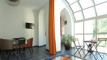 A spacious room with a large window, a flat-screen TV mounted on the wall, and a dining area with chairs and a table.