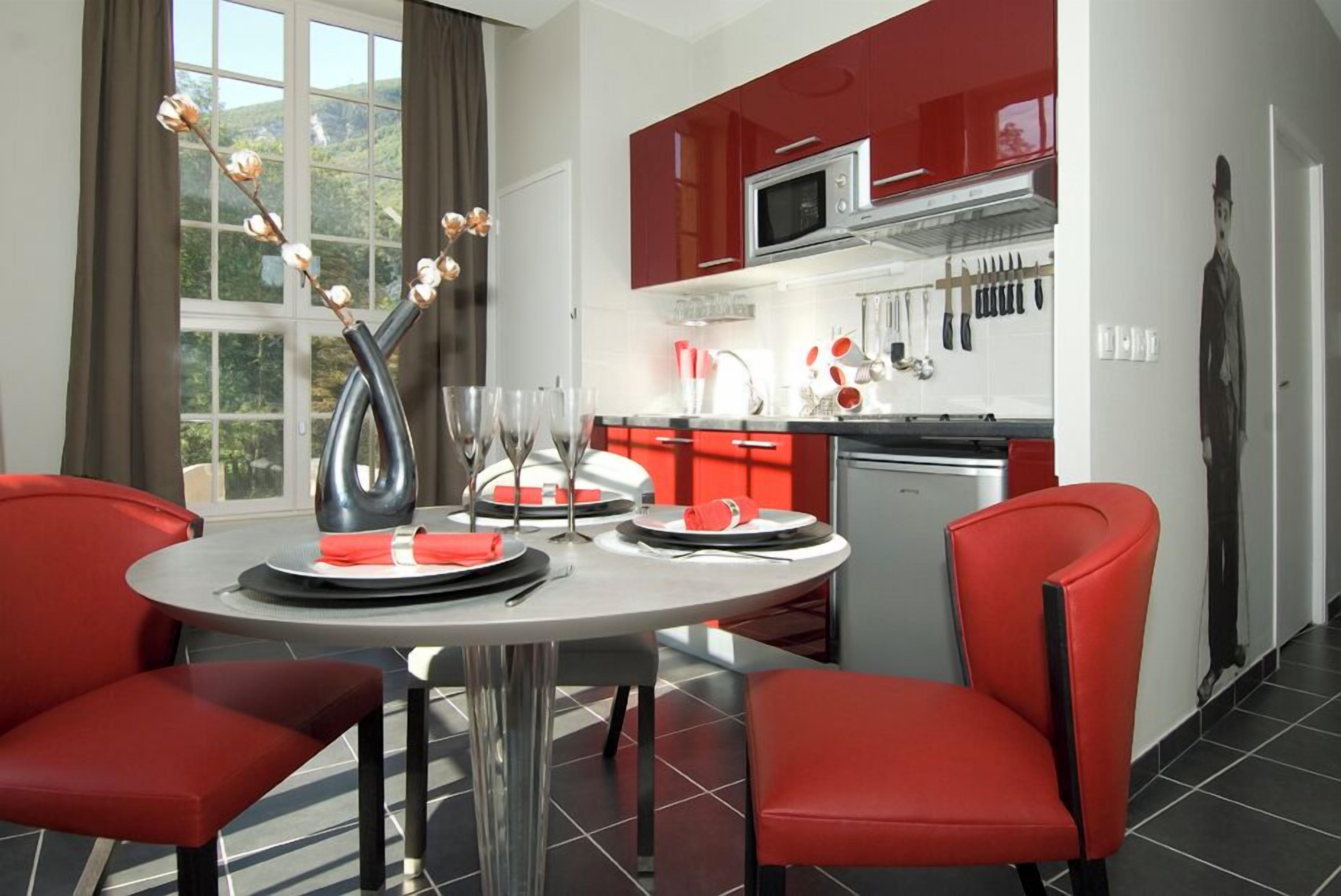 Cocina moderna con armarios rojos, microondas y lavamanos. Hay una mesa de comedor con sillas y un florero con flores.