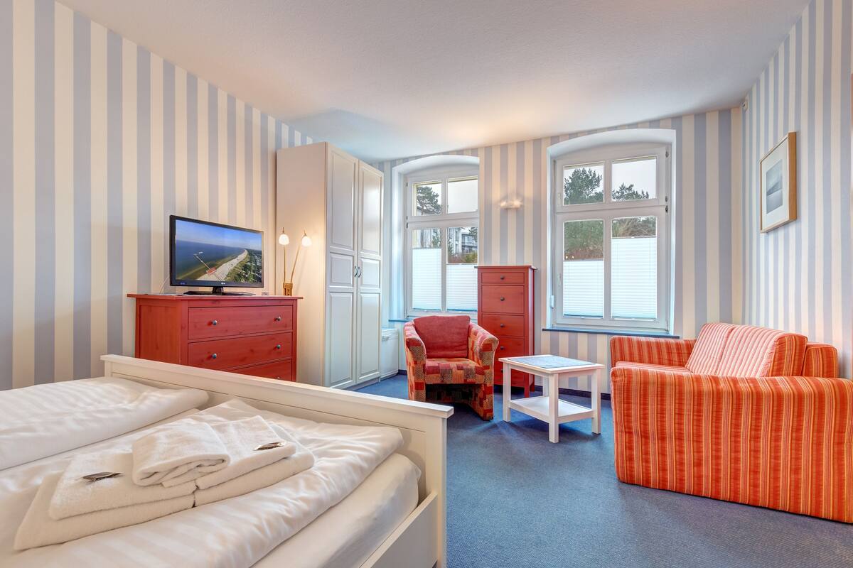 Una habitación de hotel con una cama, un sillón rojo, un sofá blanco, una mesita y un televisor sobre un mueble aparador.