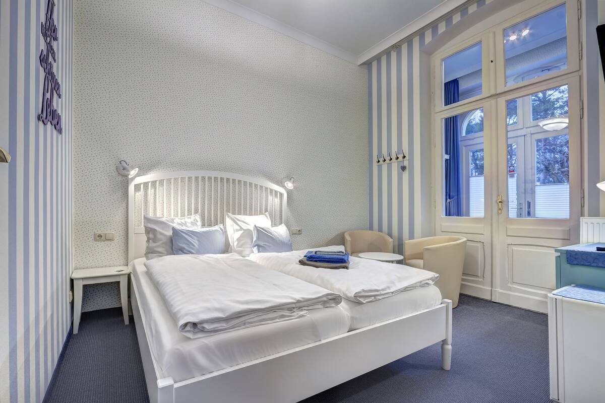 Un dormitorio ordenado con cama, mesita de noche, silla y una ventana con cortinas.