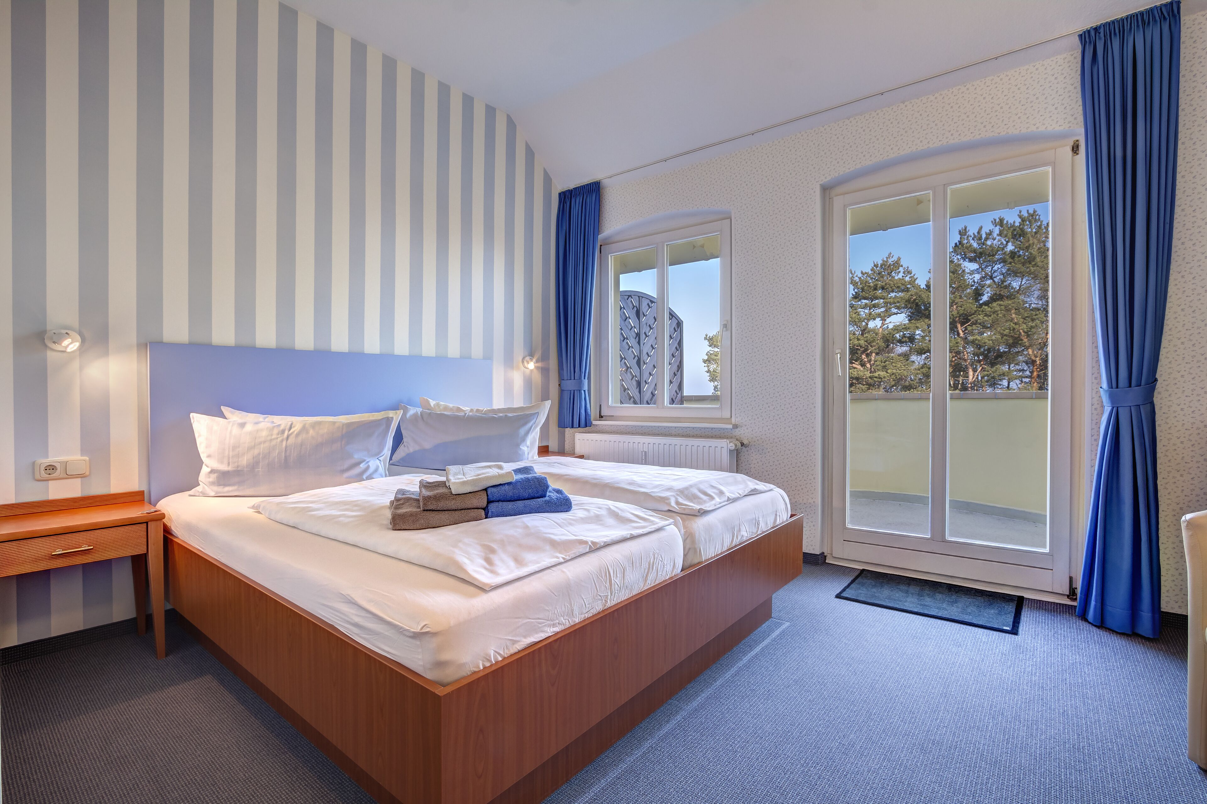 Ein Hotelzimmer mit einem großen Bett, einem Nachttisch aus Holz, einem Fenster mit Vorhängen und Blick auf Bäume.