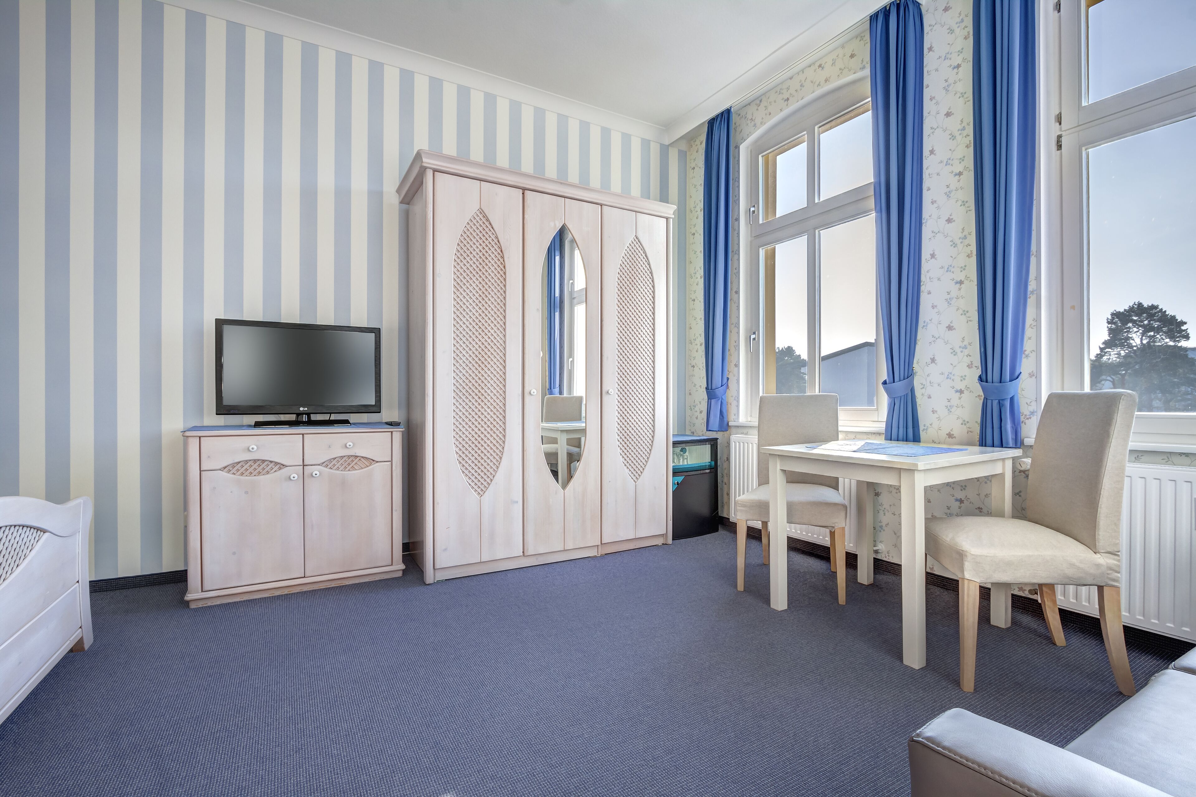 A hotel room with a striped wall, a wardrobe, a television, a dining table with chairs, and a window with curtains.
