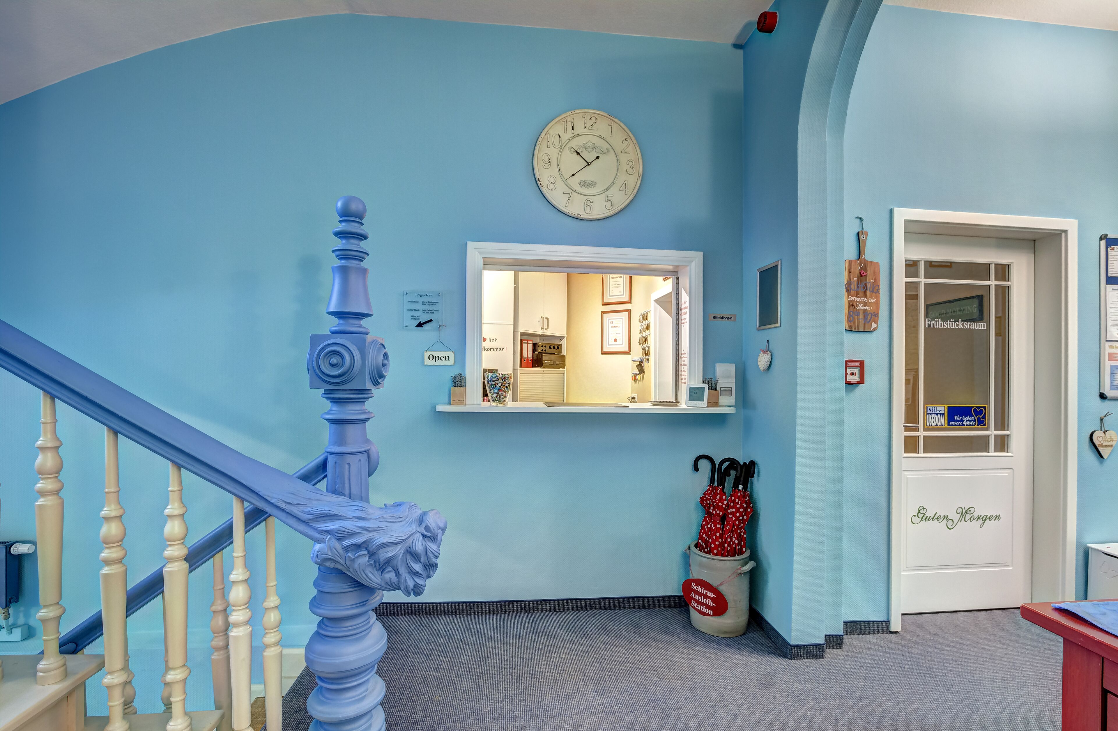 A hallway with a blue wall, a wooden staircase, a clock, a door labeled 'Frühstücksraum', and a trash bin with an umbrella.