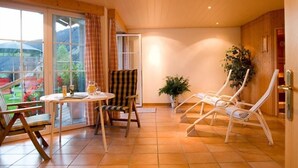 A room with a large sliding glass door leading to a garden, a small round table with a chair, and two lounge chairs. - Alpenrösle (Balderschwang)