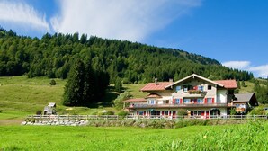 A large, two-story chalet with a red roof, surrounded by lush green fields and a forested hill. - Alpenrösle (Balderschwang)