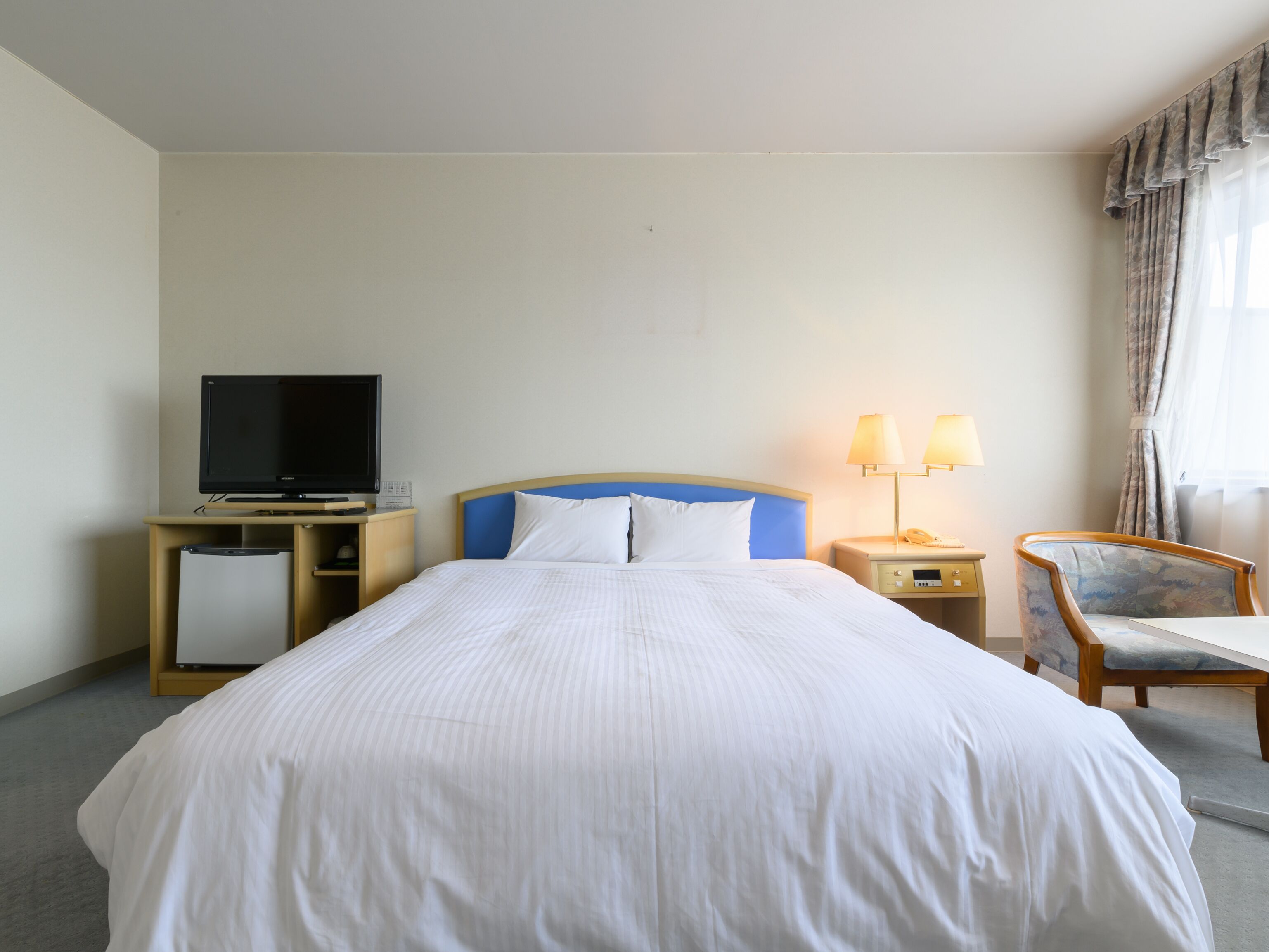 Habitación de hotel con una cama, televisor sobre un mueble, escritorio con lámpara y una silla.