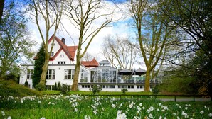 A large white building with a red roof, surrounded by trees and a field of white flowers. - Parkhotel Herne (Herne)