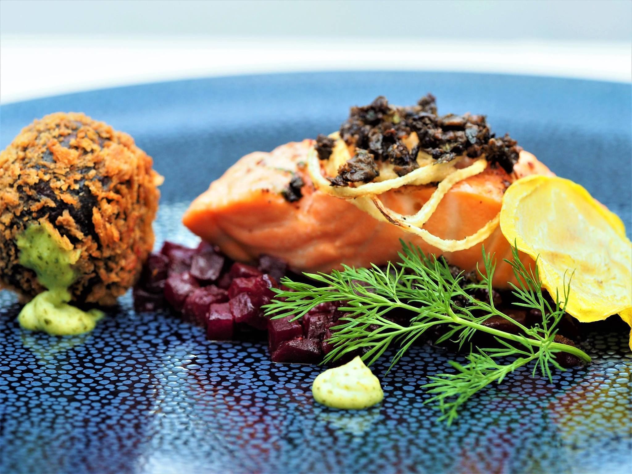 Een bord met zalm, bieten en een verkruimeld ingrediënt, met een blauwe, gestructureerde ondergrond.