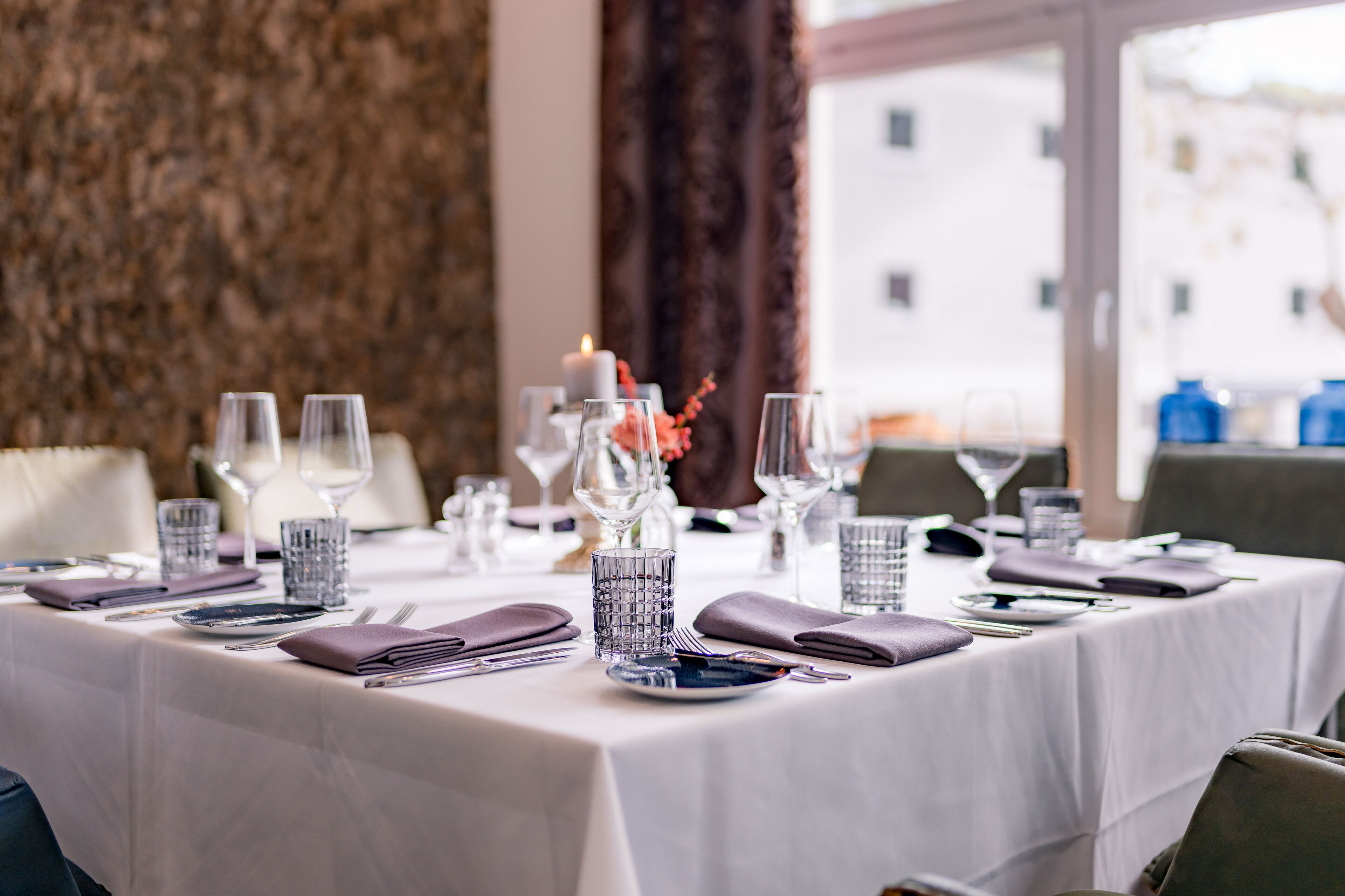 A neatly set dining table with glassware, plates, and cutlery, a candle centerpiece, and a view of buildings through a window.