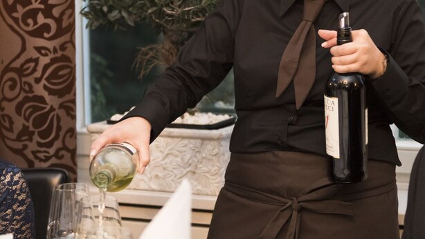A person pouring wine into a glass at a table with other glasses and a plant in the background. - Parkhotel Herne (Herne)