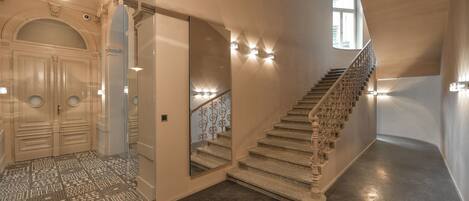 Una imponente escalera con barandillas ornamentadas y un suelo de piedra pulida, que conduce a una sala con techos altos, un espejo grande y elegantes lámparas.