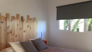 Un dormitorio con cabecera de madera, colcha a rayas, lámpara colgante blanca y ventana con persiana oscura.