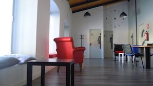 Una sala de estar moderna con un sillón rojo, una mesa negra y techo de madera. Se observan dos lámparas colgantes, un perchero y una puerta con el símbolo de silla de ruedas.