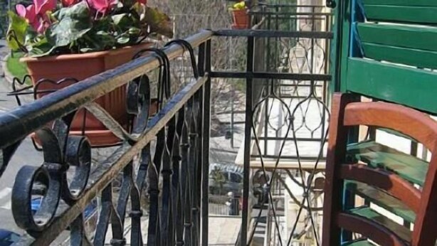 Un balcón con barandilla de metal, una silla de mimbre y una maceta con flores rojas.