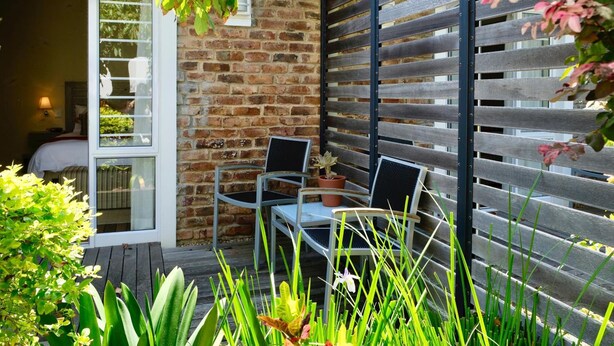 A patio area with a brick wall, a wooden bench, and two metal chairs. - Gardenview Guest House (Gqeberha (Port Elizabeth))