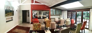 A dining area with a kitchen in the background, a red accent wall, and a view through large windows. - Gardenview Guest House (Gqeberha (Port Elizabeth))