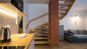 Eine moderne Küche mit einem eingebauten Ofen und einem Wasserkocher auf der Arbeitsplatte. Eine Ziegelstütze trägt eine Holztreppe, die zu einer anderen Ebene führt. Ein graues Sofa ist im Hintergrund zu sehen.