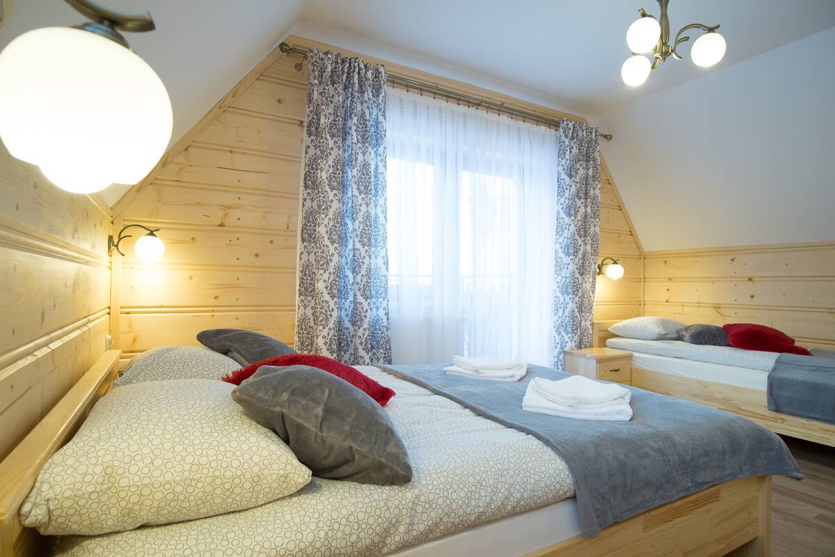 Ein gemütliches Schlafzimmer mit Holzwänden, einem großen Fenster mit gemusterten Vorhängen und zwei Betten mit ordentlich bezogenen Laken.