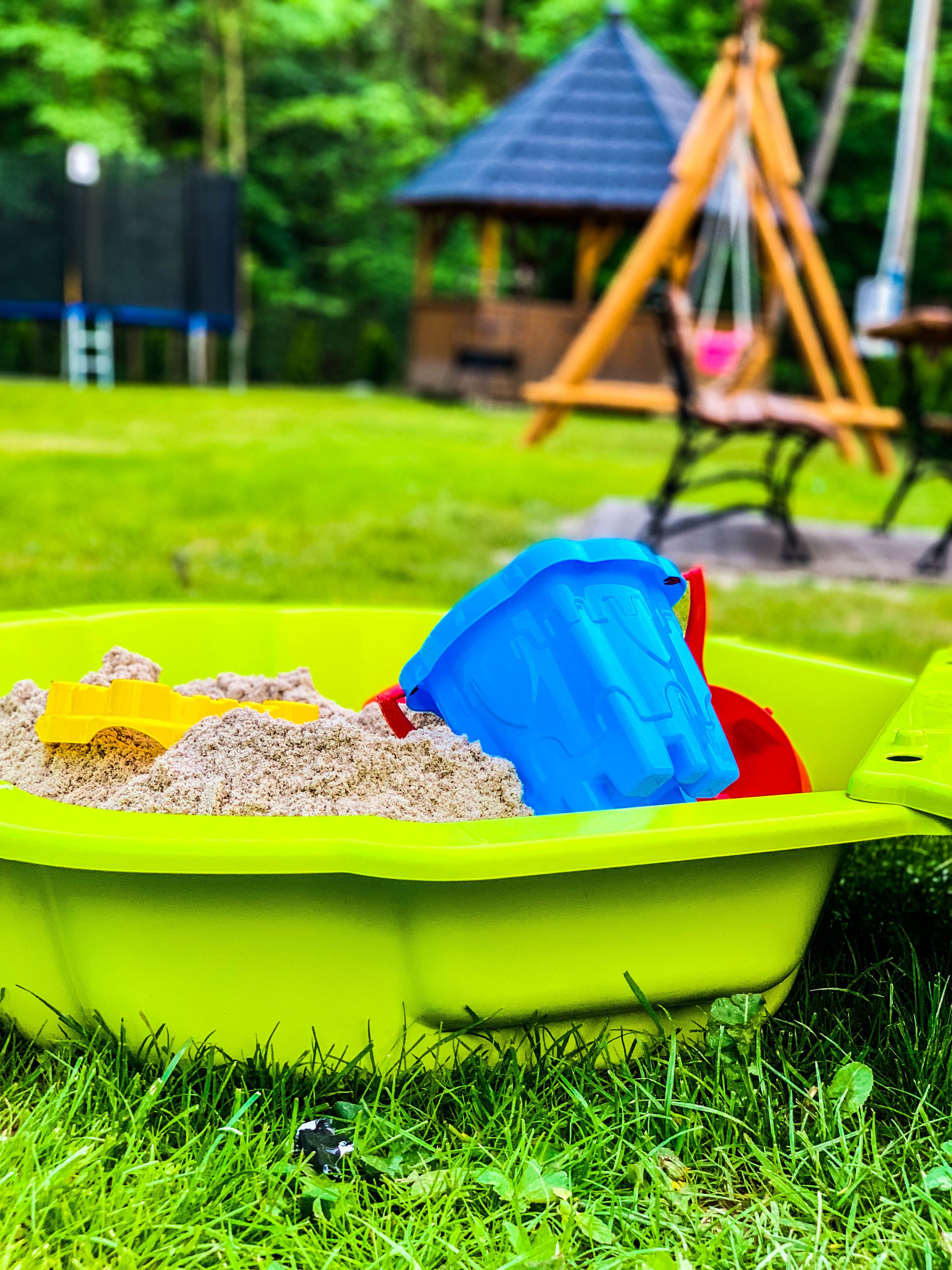 A green plastic container filled with sand and a blue shovel on a grassy lawn with a swing set and a gazebo in the background.