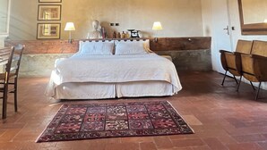 Une chambre avec un grand lit, des poutres apparentes en bois au plafond, un tapis à motifs et un fauteuil.