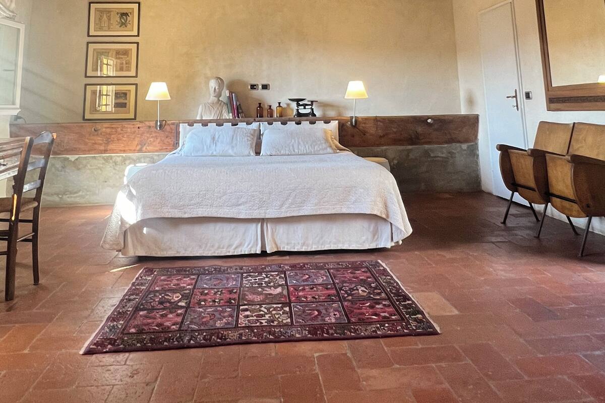 Een slaapkamer met een groot bed, houten balken in het plafond, een kamerbreed tapijt met dessin en een stoel.