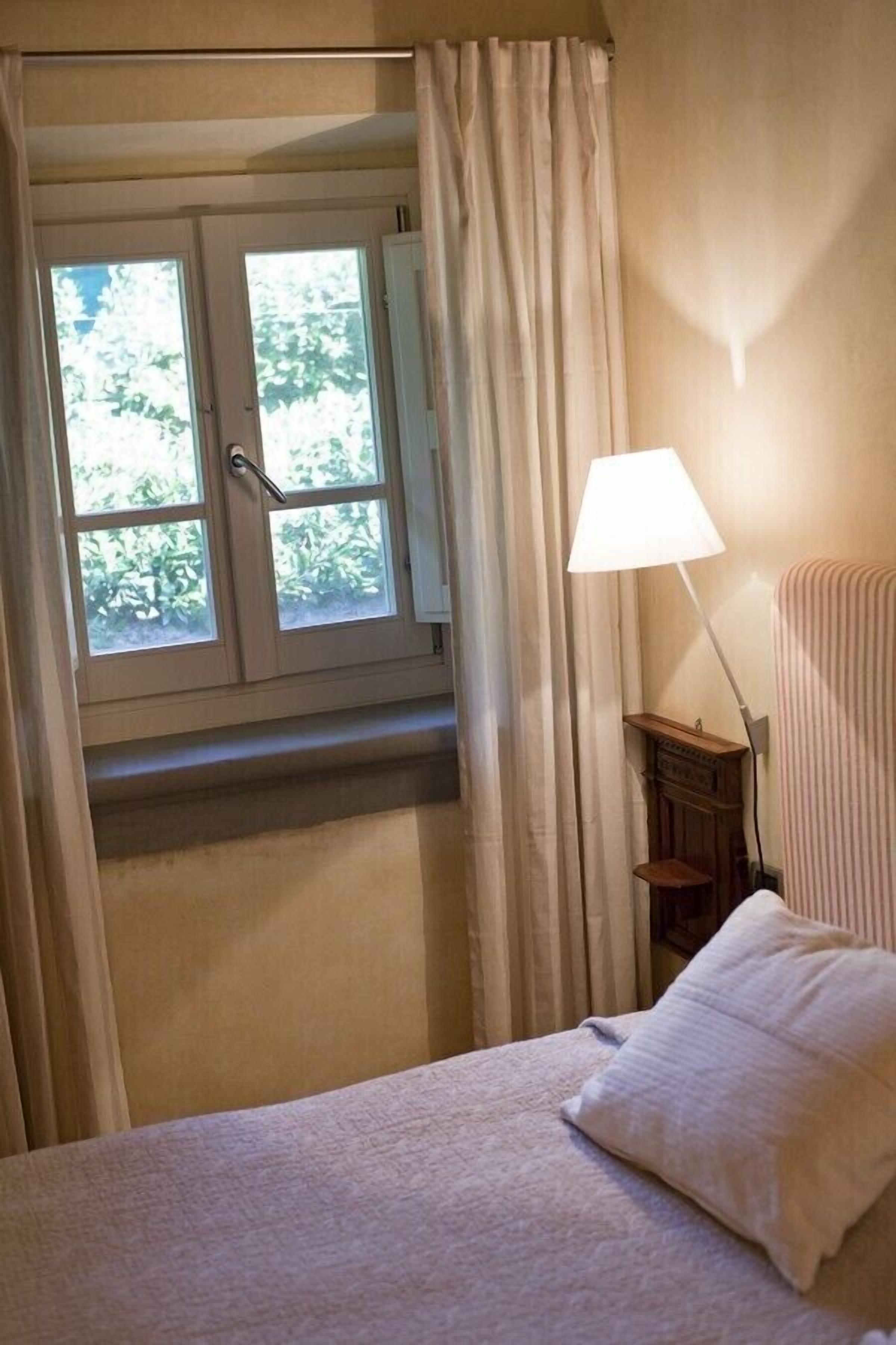 Un dormitorio con cama, ventana con cortinas, lámpara de noche y mesita de noche.