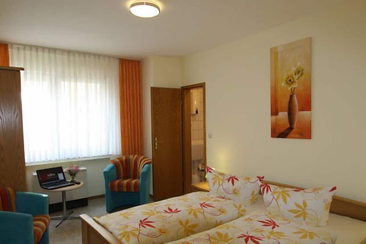 A hotel room with a bed, two chairs, a desk with a laptop, a painting on the wall, and a lamp.