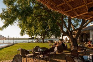 Un área de comedor al aire libre con mesas y sillas bajo techo de paja, con vista a un cuerpo de agua y un cartel de Coca-Cola al fondo.