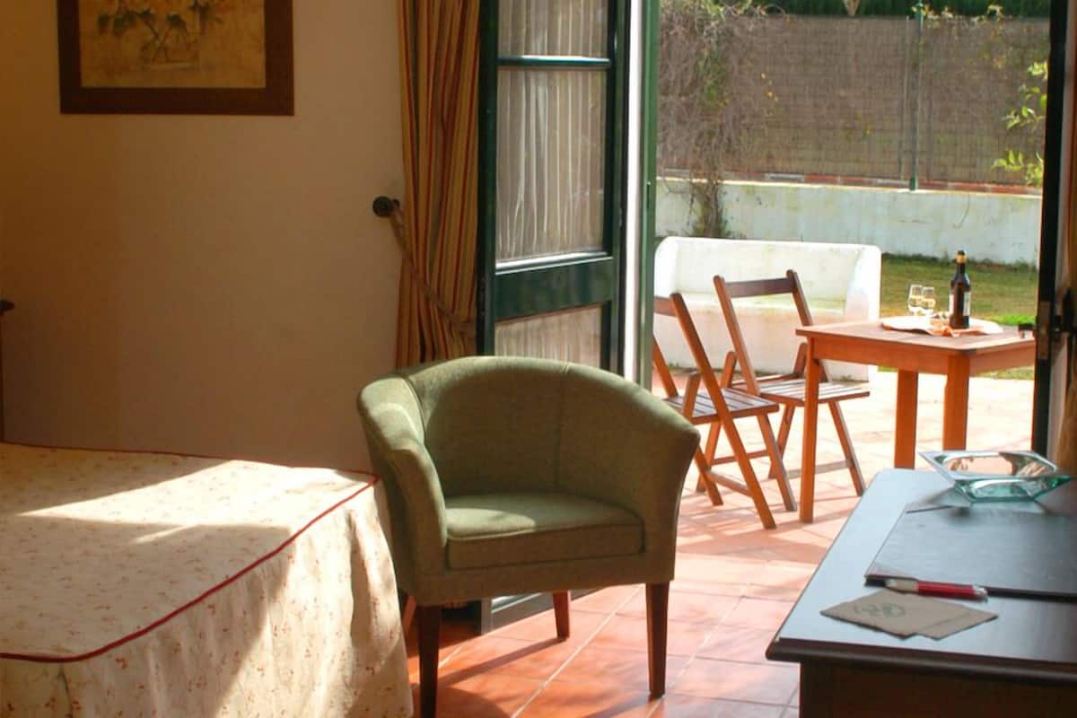 Une pièce avec un lit, un fauteuil vert, une table en bois sur laquelle se trouvent une bouteille et des verres, et une vue sur un jardin par une porte ouverte.