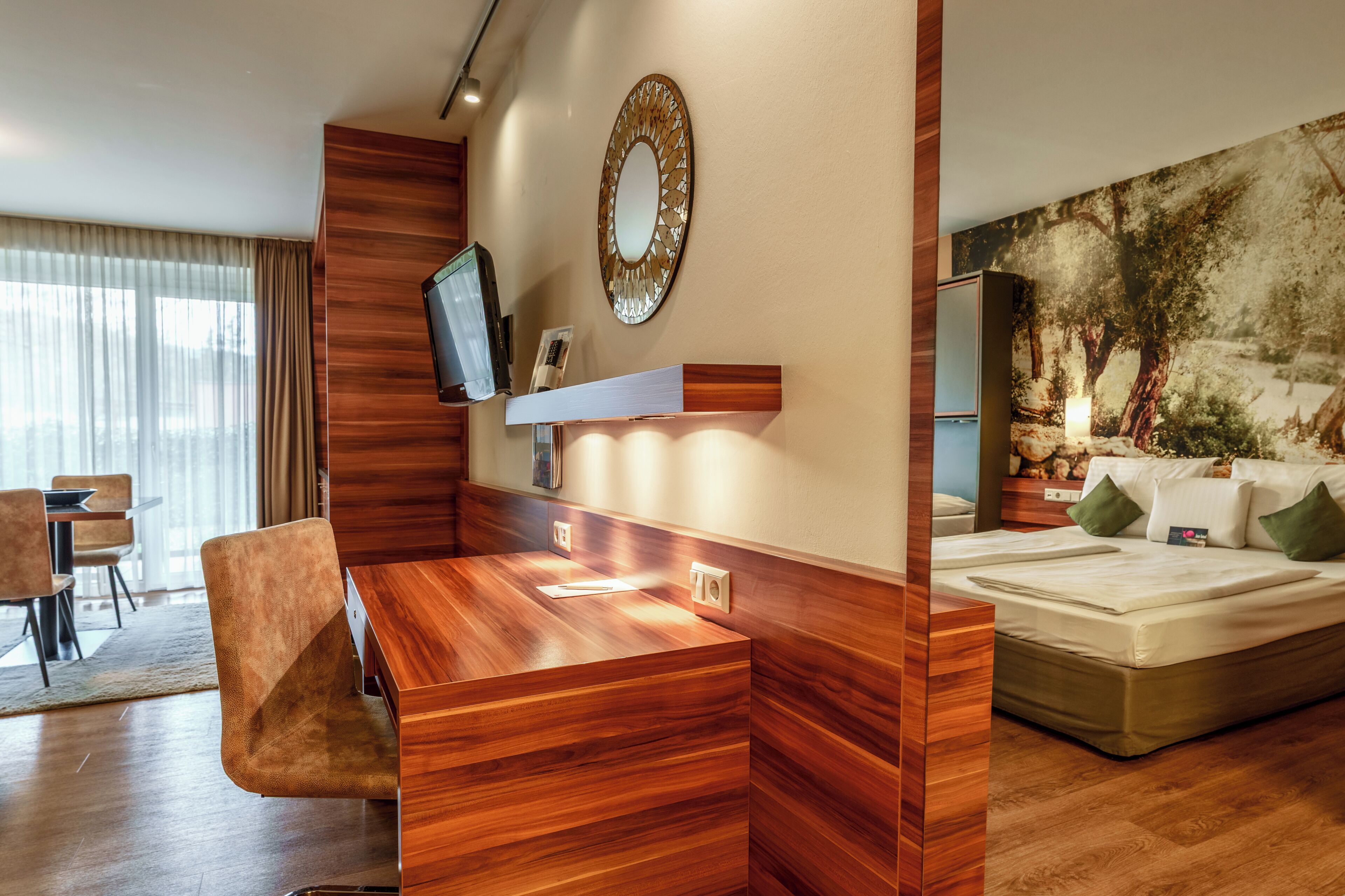 A modern hotel room with a wooden desk, a television, a large bed, and a painting on the wall.