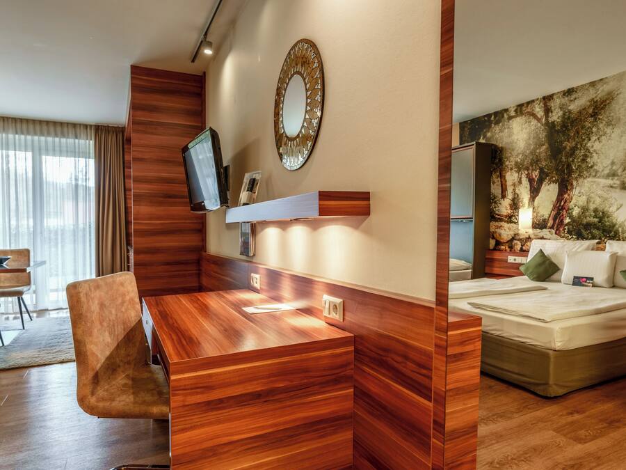A modern hotel room with a wooden desk, a television, a large bed, and a painting on the wall.