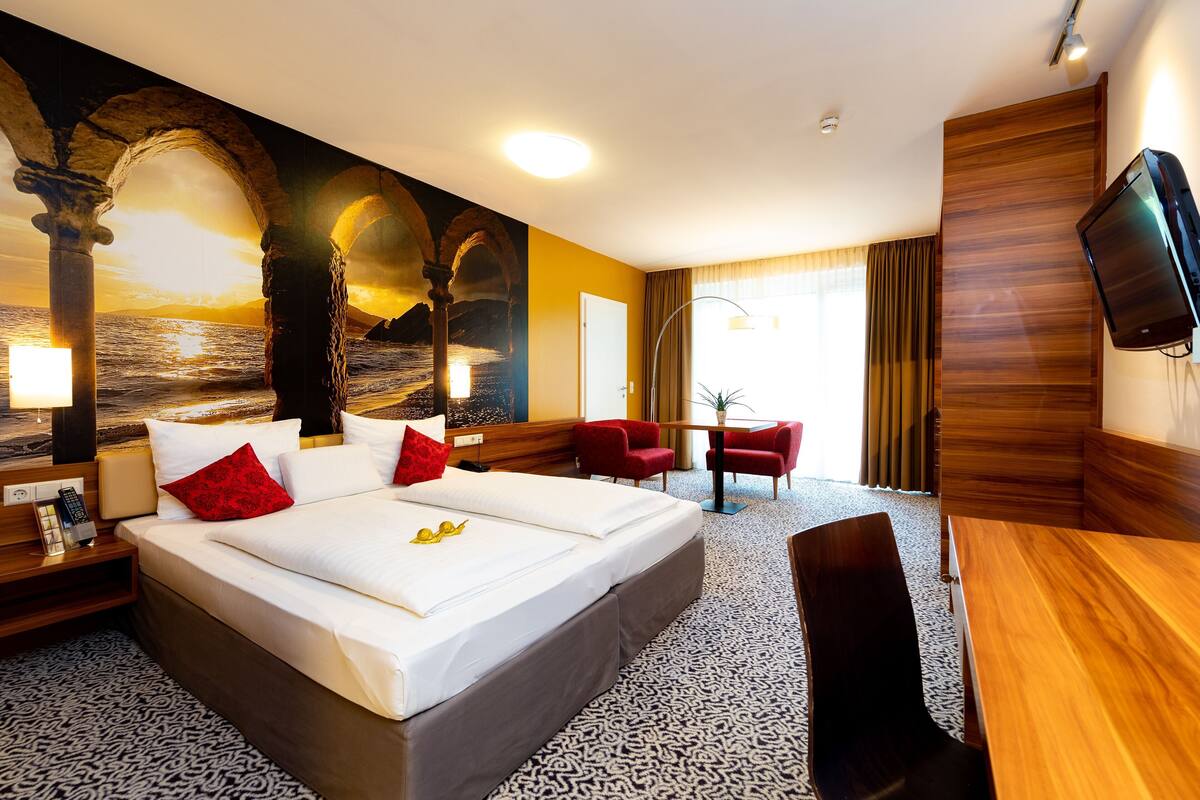 A hotel room with a large bed, a desk, a chair, a TV, and a mural of a sunset over the ocean.