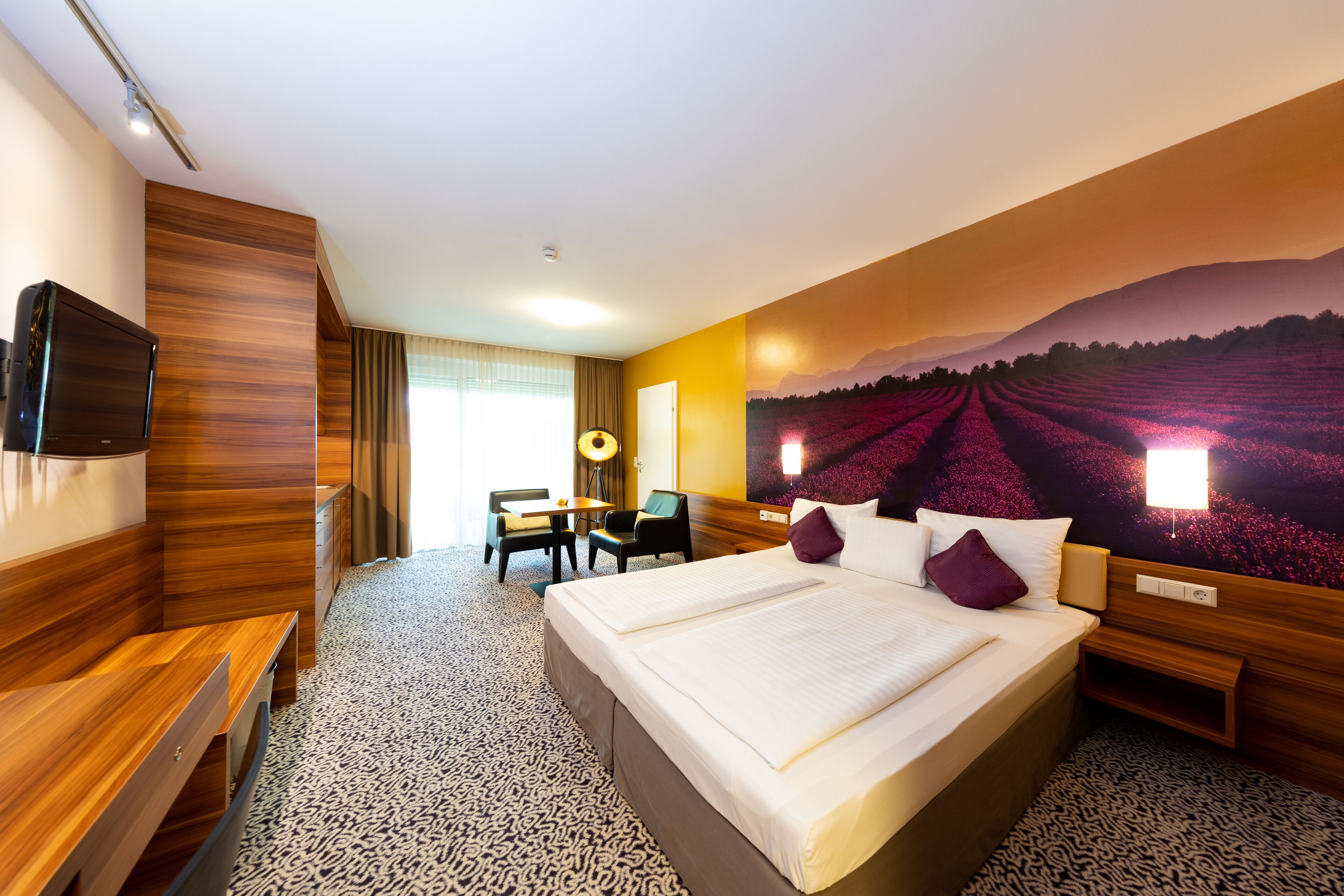 A hotel room with a large bed, a desk, and a chair. There is a television on the wall and a painting of a field on the wall.