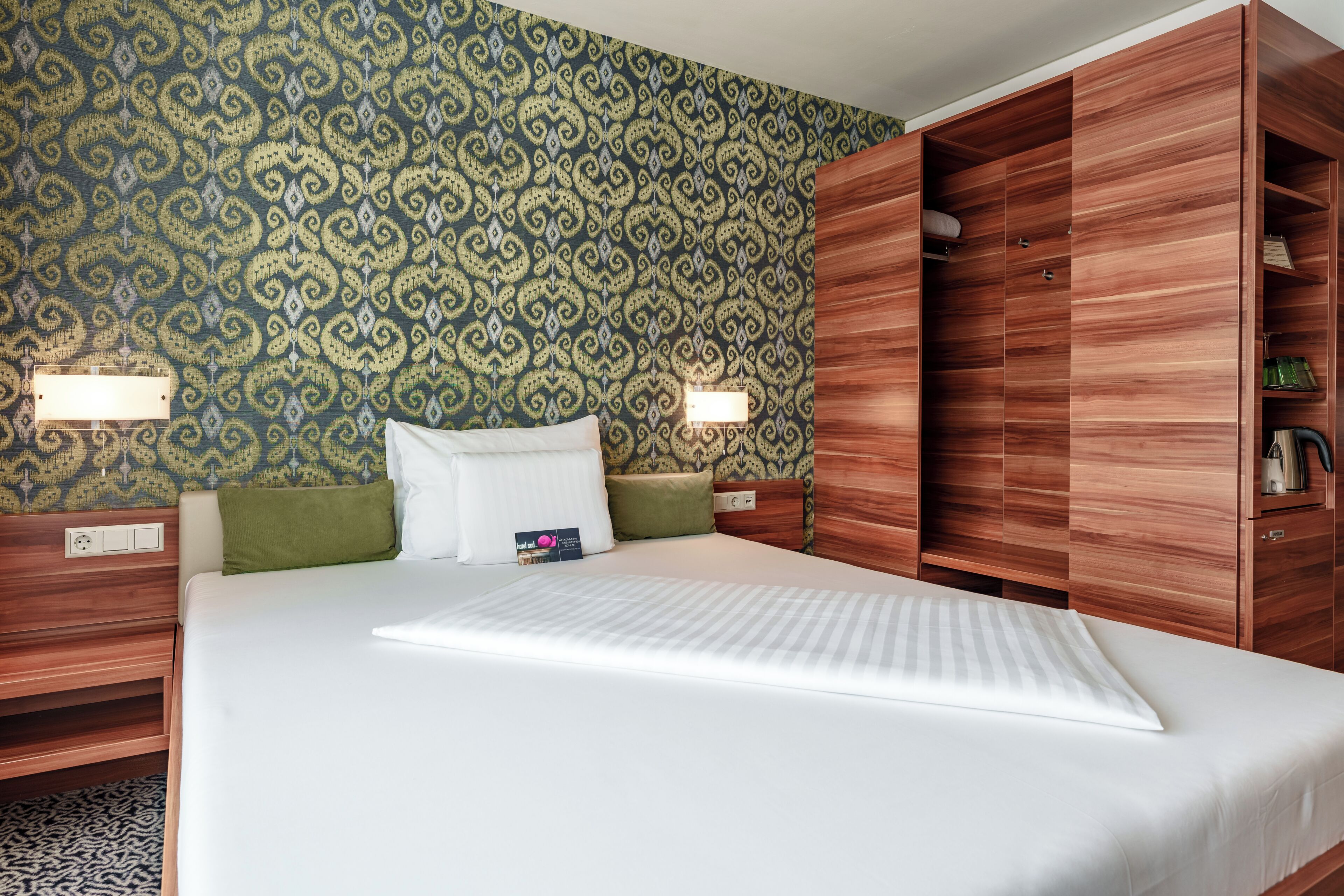 A neatly made bed with a white mattress and pillows, a wooden wardrobe with a mirror, and a patterned wallpaper.