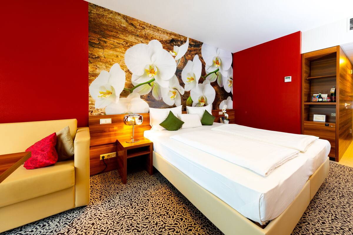 A hotel room with a bed, a nightstand, a lamp, a red wall, and a wooden cabinet.