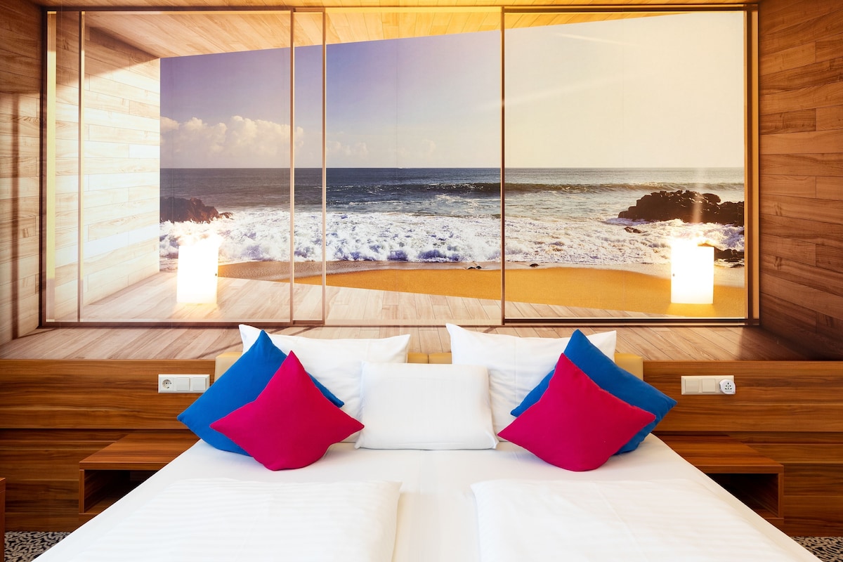 A hotel room with a bed, pillows, and a large window with a beach view.