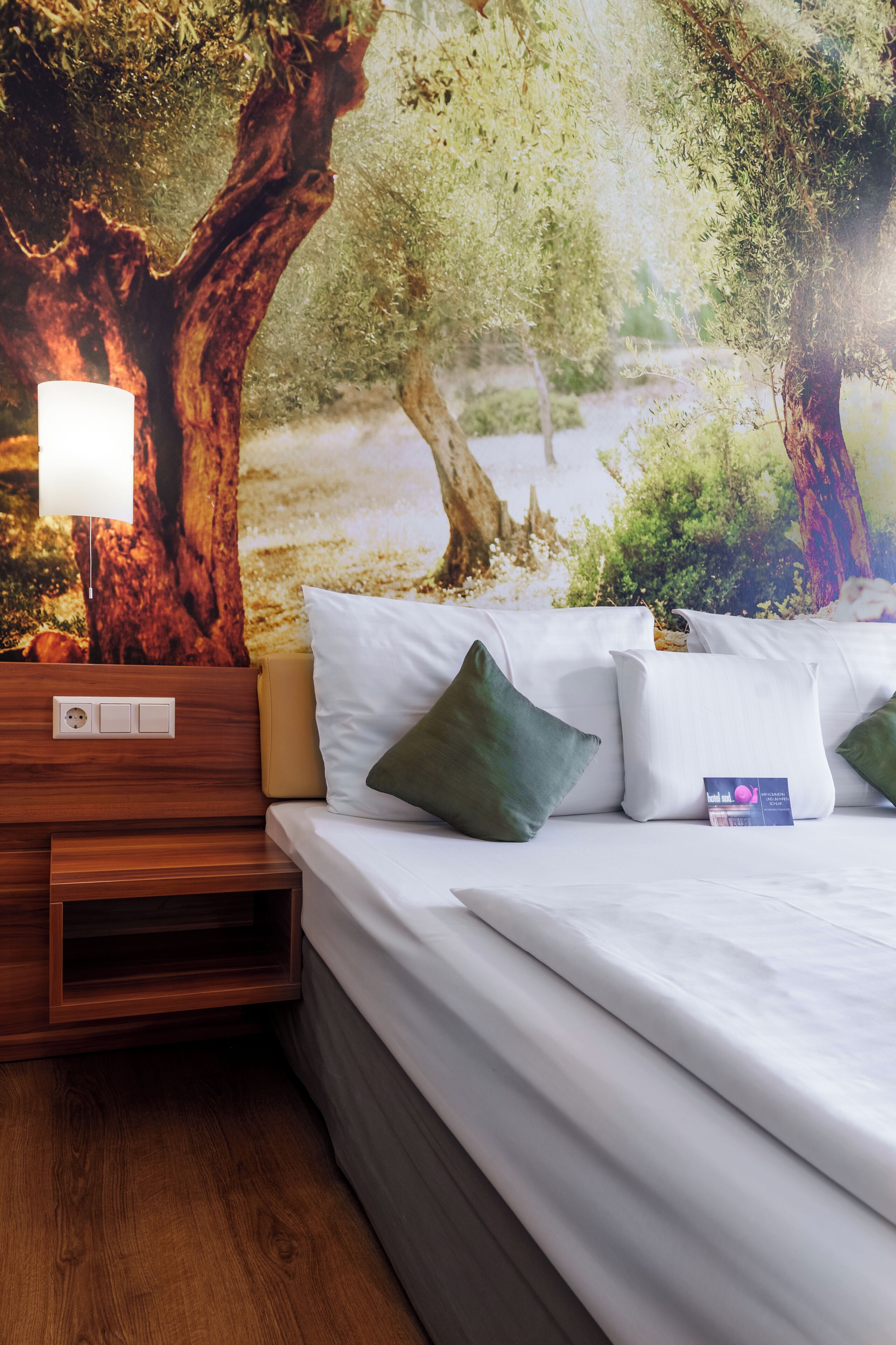 Habitación de hotel con cama, almohadas, mesita de noche, lámpara y un mural de árboles en la pared.
