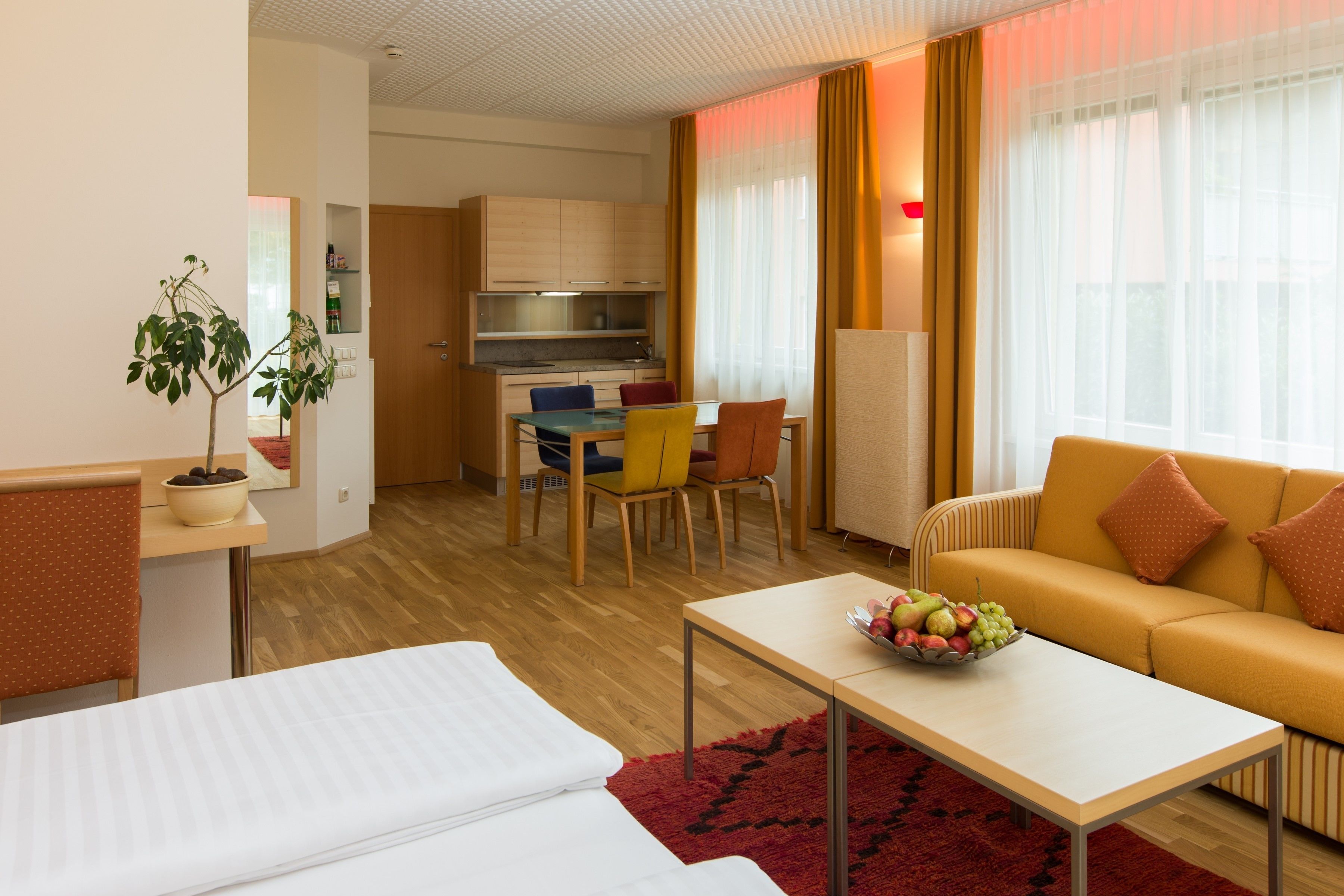 A hotel room with a bed, a dining table with chairs, a sofa, and a kitchen area.