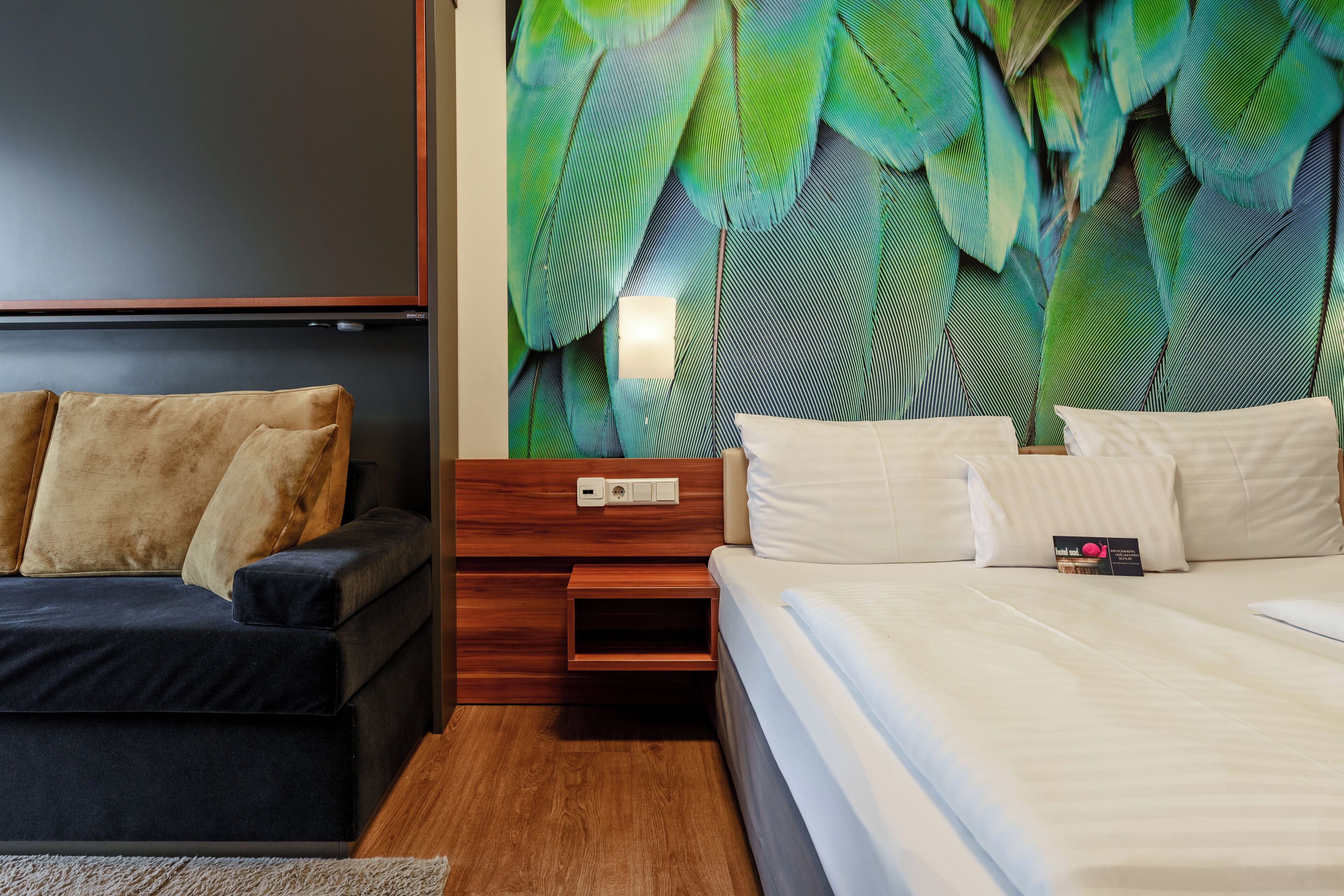 A modern bedroom with a dark sofa, a wooden nightstand, a bed with white linens, and a wall with large green leaves.