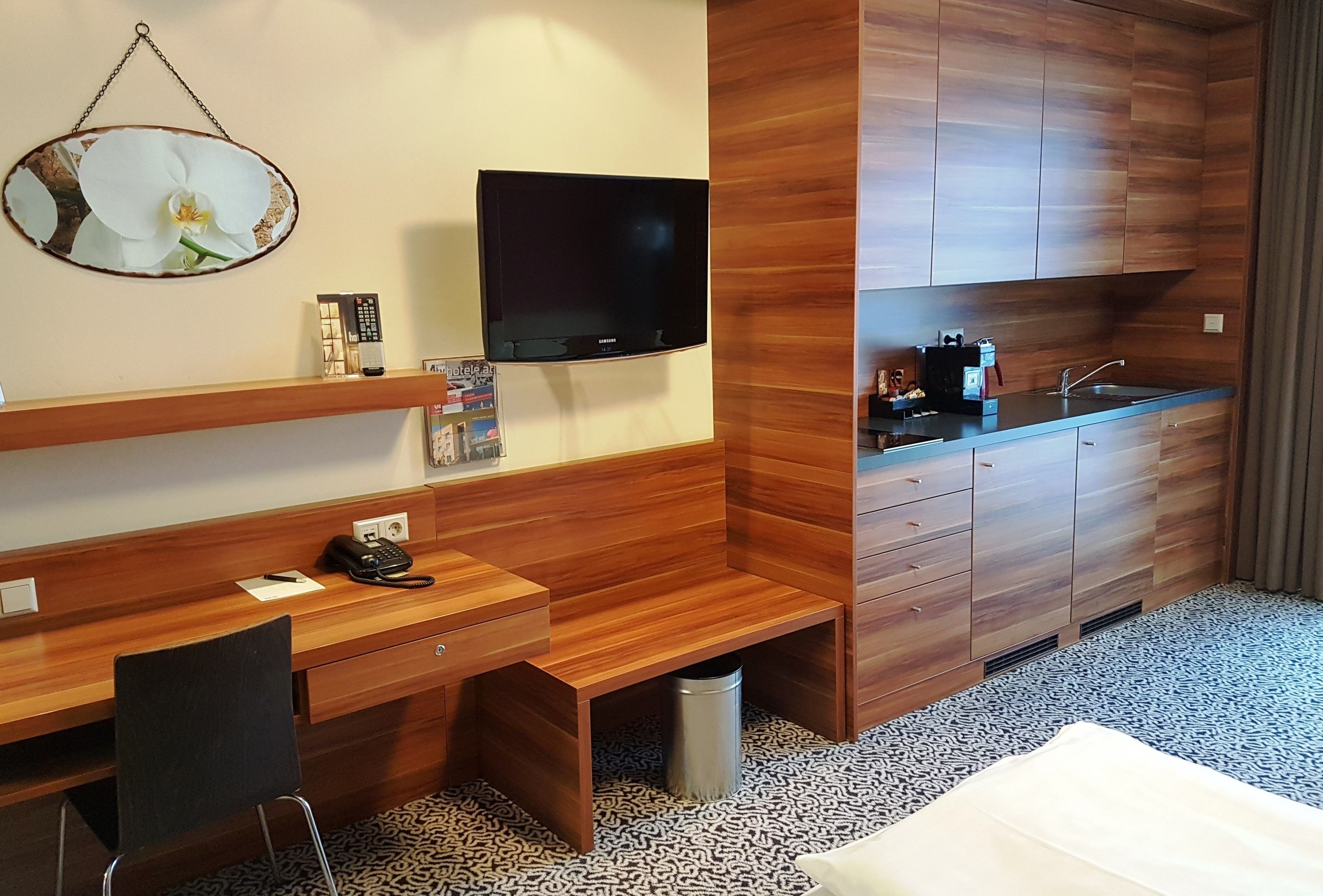 A hotel room with a wooden desk, a television, a small kitchenette, and a bed.