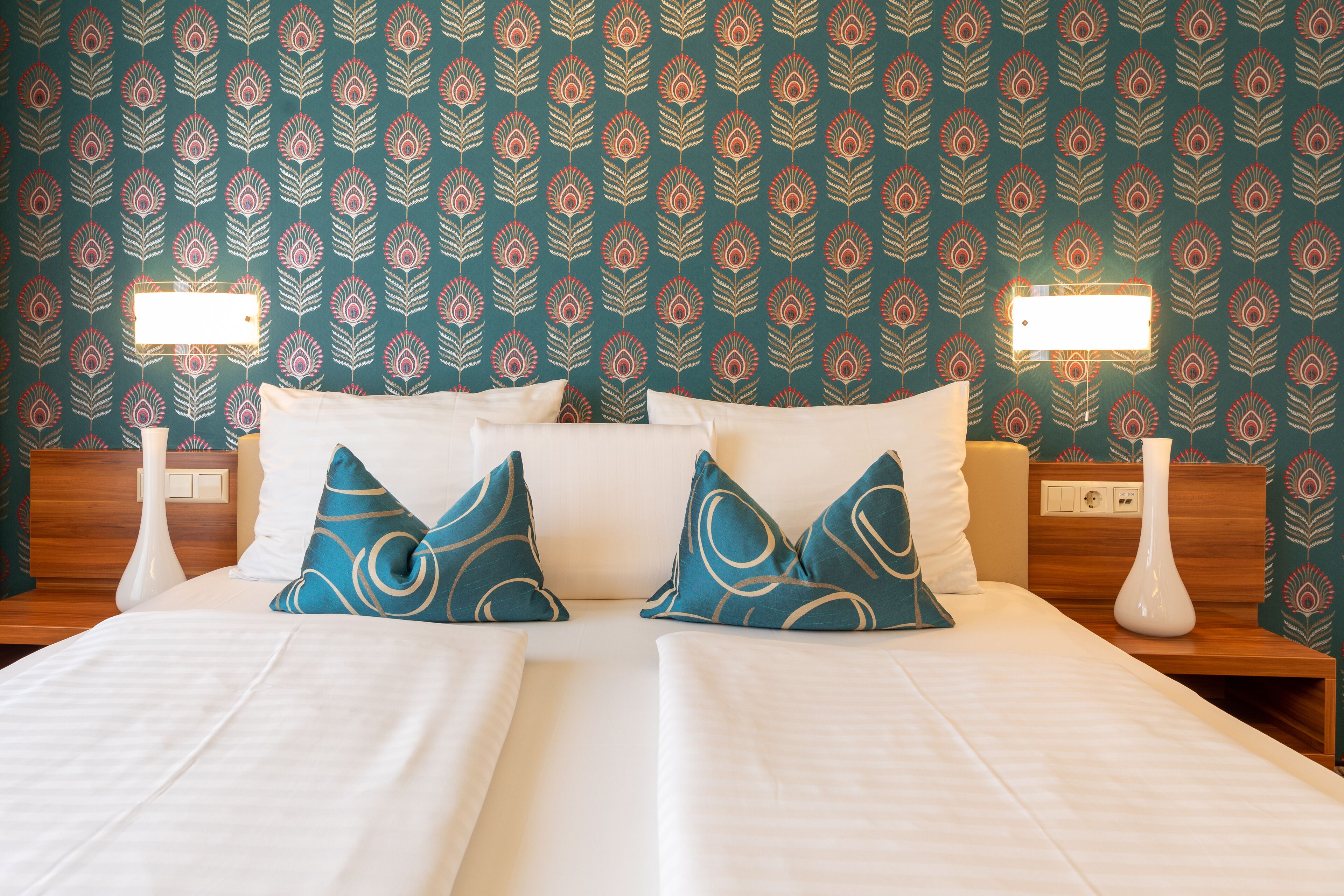 Una cama doble con ropa de cama blanca y dos almohadas decorativas, una mesita de noche con un florero blanco, y una pared con papel tapiz estampado.