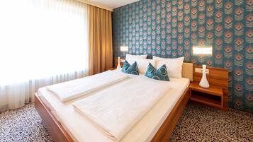 Cama de casal com lençóis brancos, cabeceira de madeira e almofadas decorativas. Há uma mesa de cabeceira com um vaso branco e um abajur. O quarto tem papel de parede estampado e uma janela com cortinas.