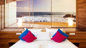 Hotelkamer met een groot uitzicht op het strand door een glazen schuifdeur, een bed met witte en roze kussens en houten lambrisering.