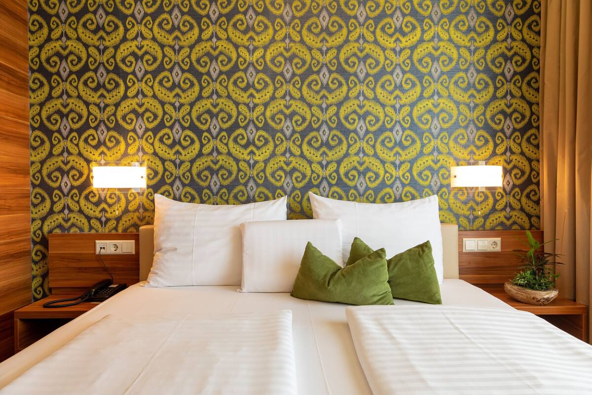 A hotel room with a patterned wallpaper, a bed with white linens, two bedside lamps, a bedside phone, and a nightstand with a plant.