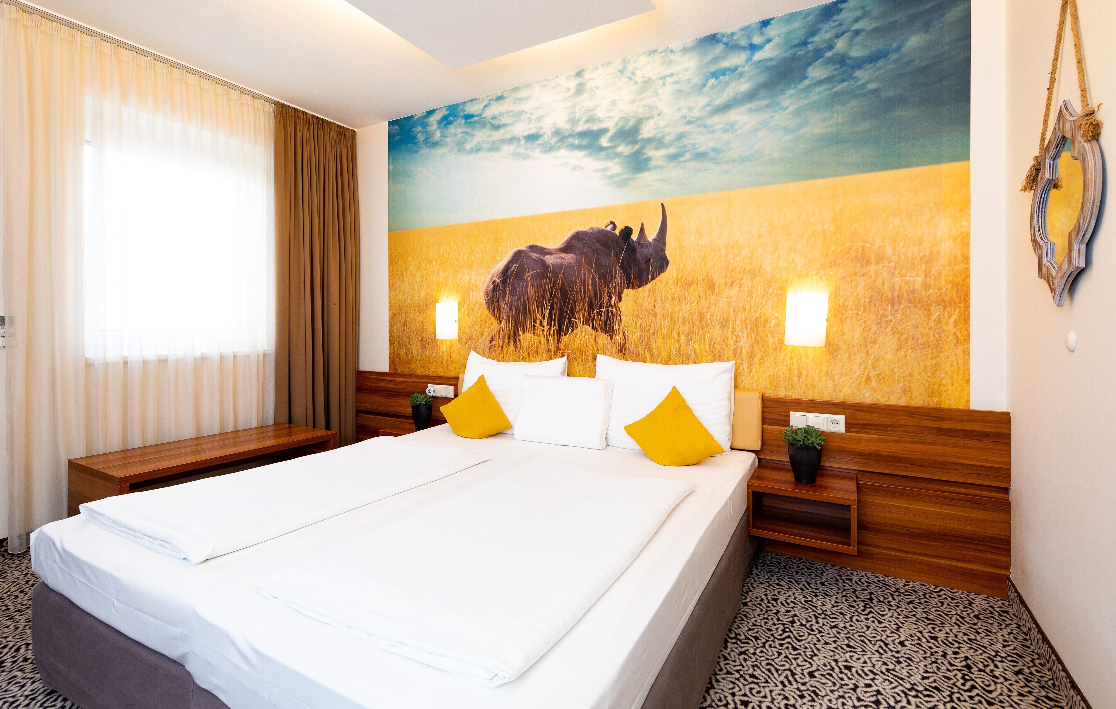 Habitación de hotel con una cama grande, cabecera de madera, dos mesitas de noche, un mural en la pared con un rinoceronte y una ventana con cortinas.