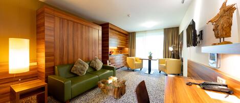 Habitación de hotel moderna con sofá verde, paneles de pared de madera, alfombra estampada, una mesa de comedor de madera y una mesita auxiliar.