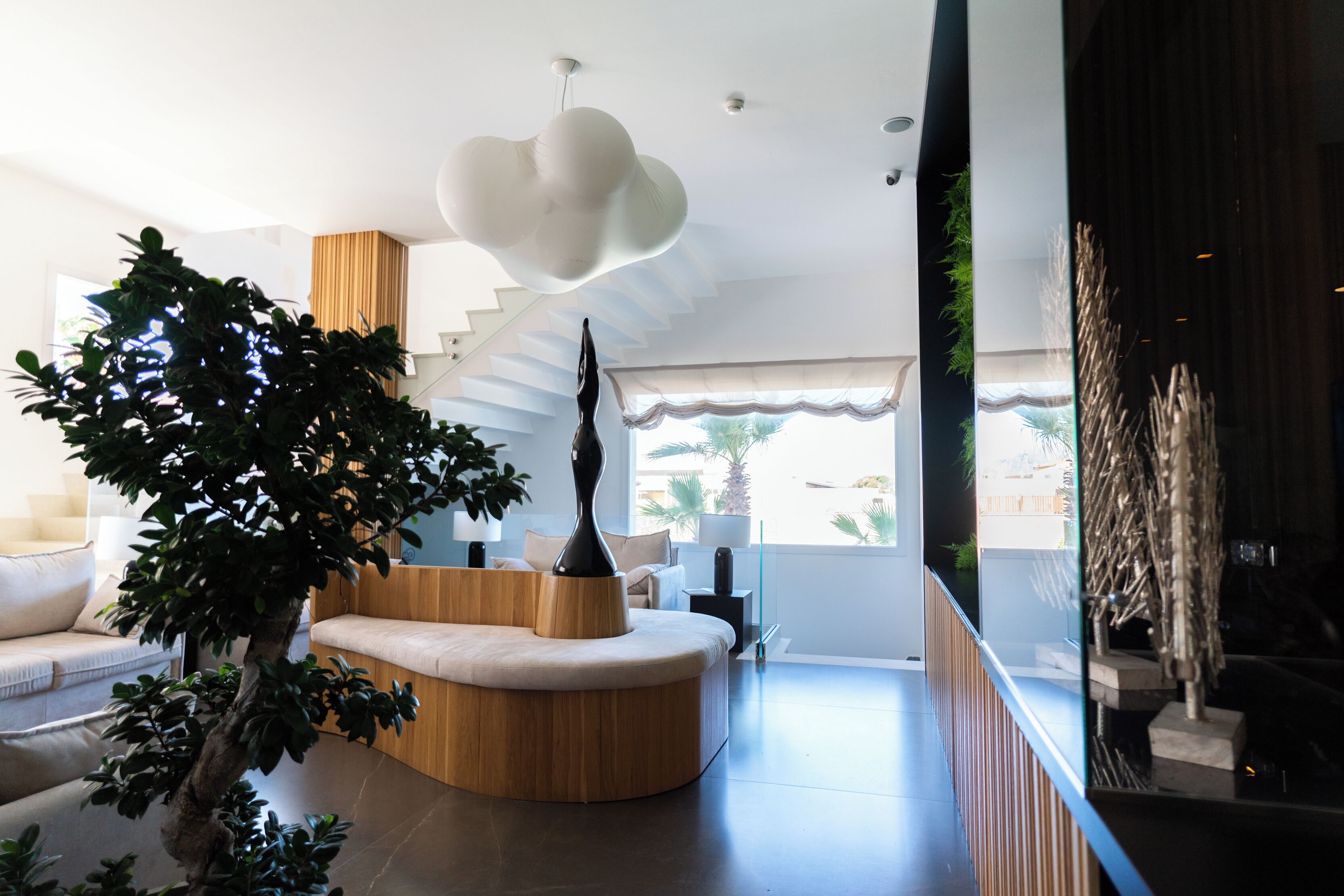 A modern living room with a curved wooden sofa, a tall black sculpture, and a large white pendant light.