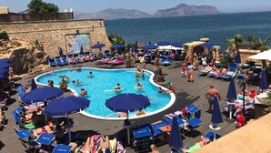 A pool area with lounge chairs and umbrellas, surrounded by a stone wall and a view of the sea.