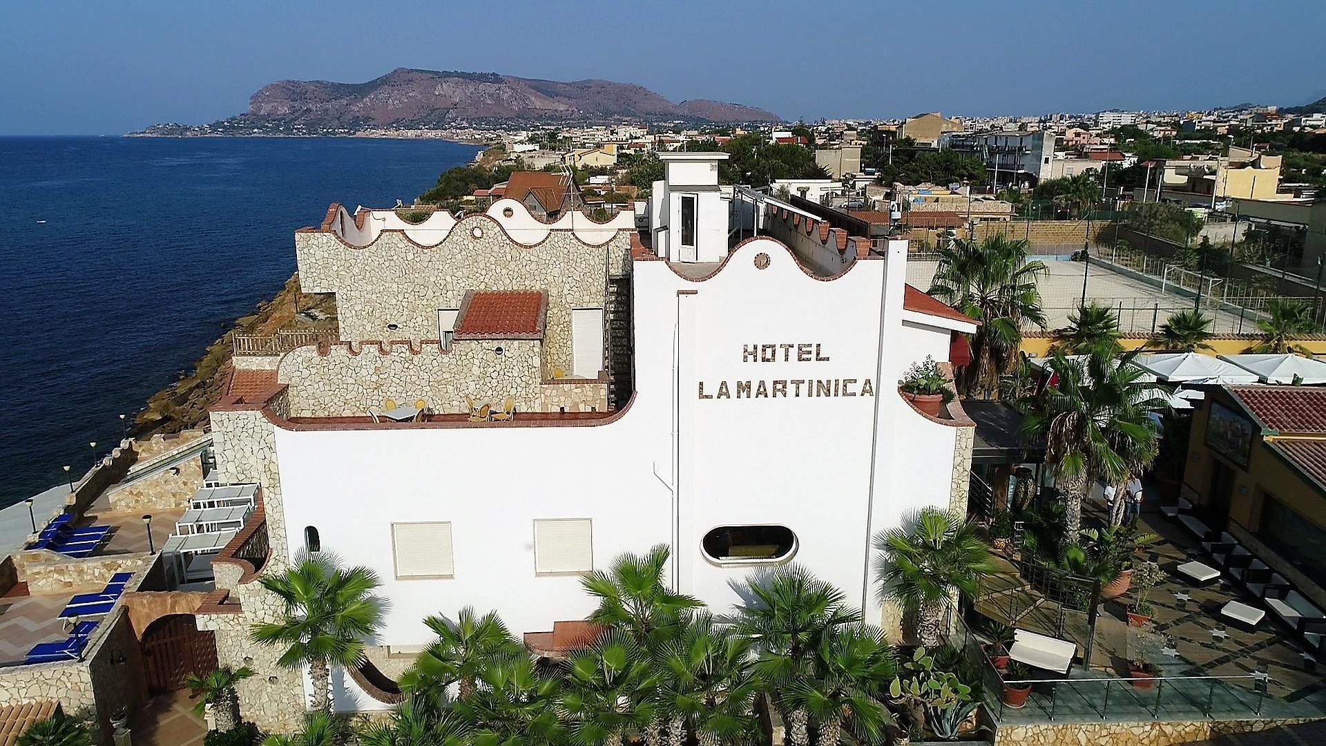 A white building with "HOTEL LAMARTINICA" on the facade, surrounded by palm trees and a view of the sea.